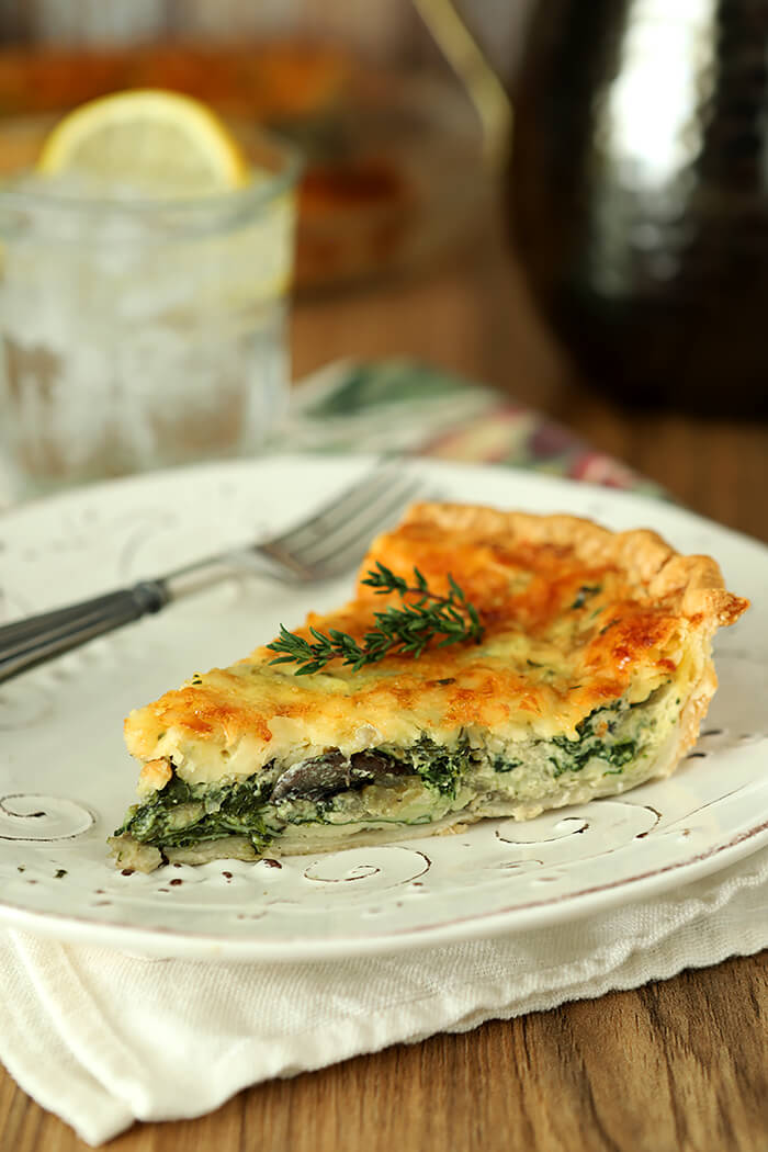 Spinach, Onion, and Mushroom Quiche with White Cheddar Cheese Sliced and Served on a White Plate with Thyme Garnish