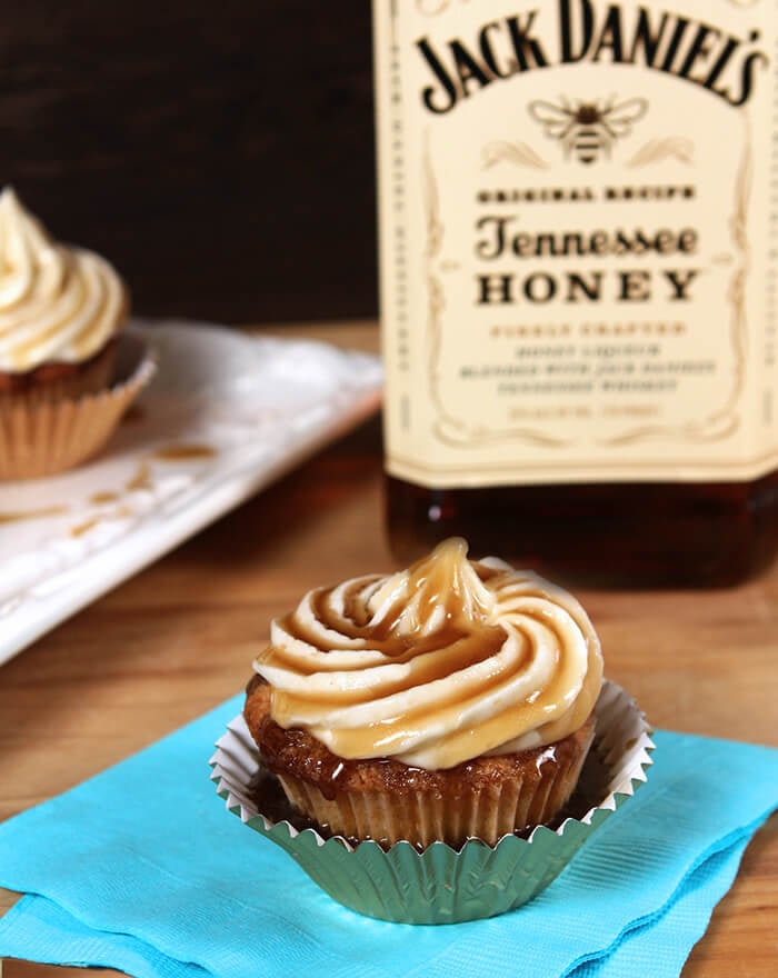 Closeup of Jack Daniels Honey Whiskey Cupcakes with a Boozy Drizzle from a Spoon In a Metallic Cupcake Liner in Front of a Bottle of Jack Daniels Tennessee Honey