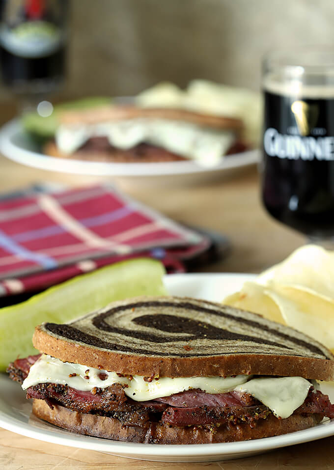Home Cured Pastrami on Rye Sandwiches with Swiss Cheesee and Whole Mustard Home Cured Pastrami on Rye Sandwiches with Swiss Cheese served on a plate; Guinness beer in the background.