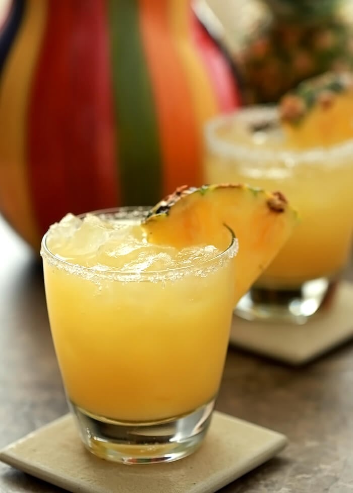 Fresh Pineapple Margarita Cocktail Served in a lowball glass with a multi-colored pitcher in background.