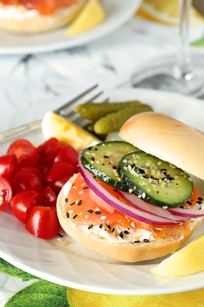 Bagels Stacked with Smoked Salmon, Cream Cheese, Red Onion and Cucumber with Tomatoes and Pickles on the Side on a White Plate with Lemon Garnish